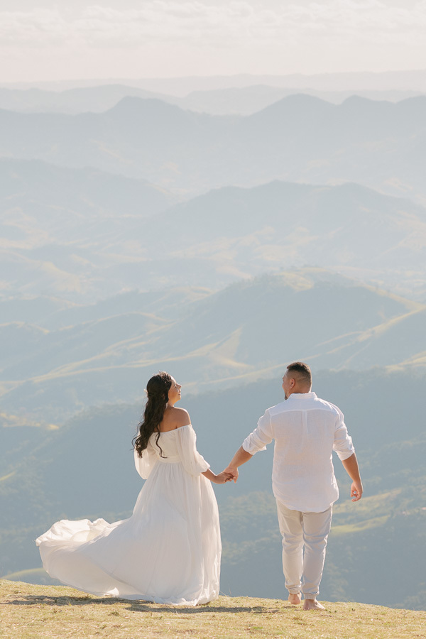 Letícia e Juliano caminham de mãos dadas, de costas, em direção ao horizonte na Pedra do Baú. Ela veste vestido branco fluido e ele camisa e calça claras. Ao fundo, as montanhas da Mantiqueira se perdem em tons de azul sob o céu claro.
