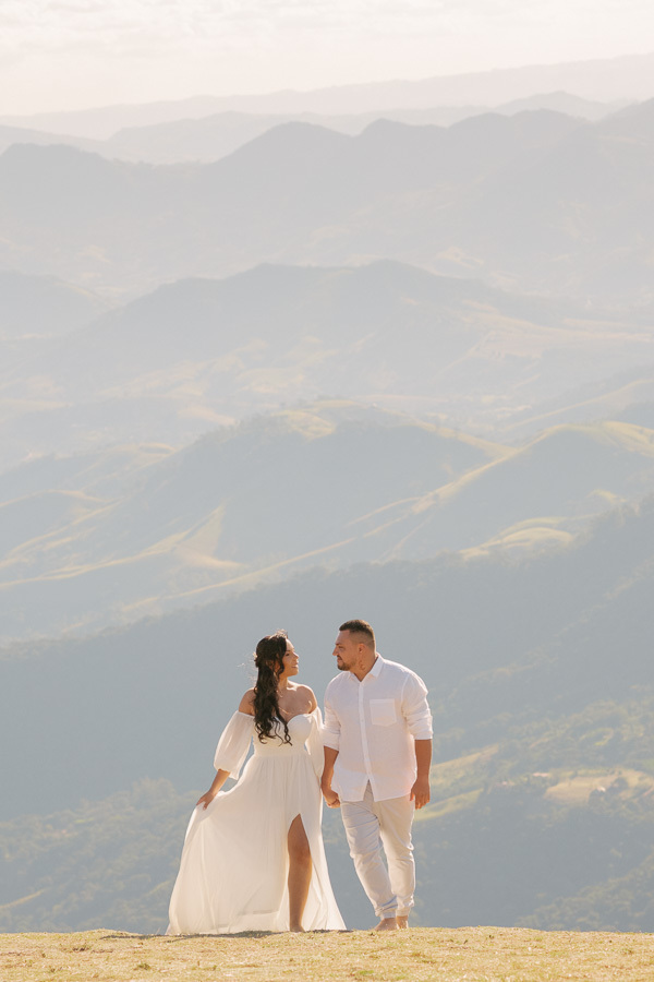 Letícia e Juliano caminham de mãos dadas no topo da Pedra do Baú, trocando olhares. Ela usa um vestido branco fluido com fenda e ele camisa e calça brancas. Ao fundo, as montanhas da Mantiqueira se estendem sob uma luz suave e céu claro.