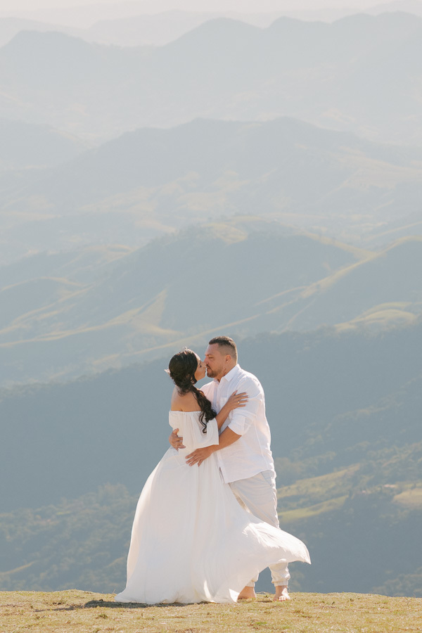 Leticia e Juliano se beijam apaixonados no topo da Pedra do Baú. Ela veste um vestido branco fluido que esvoaça com o vento e ele uma camisa branca. Ao fundo, a vista panorâmica das montanhas da Mantiqueira sob um céu claro e iluminado.