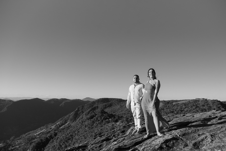 Foto em preto e branco de Letícia e Juliano no topo de uma encosta rochosa na Pedra do Baú. Ela veste um vestido justo de um ombro só com fenda; ele usa camisa e calça claras. Ao fundo, a vista profunda dos vales e montanhas da Serra da Mantiqueira sob um