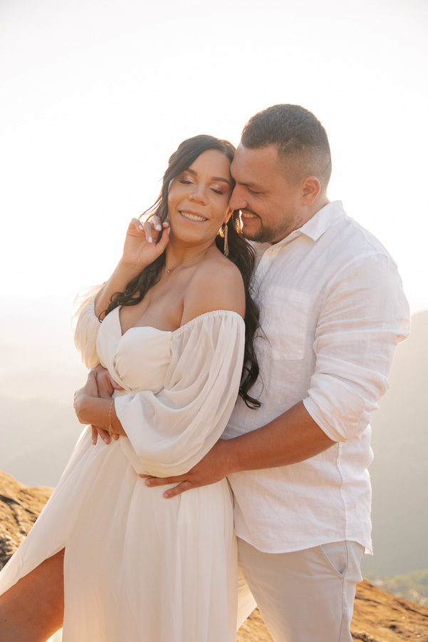 Juliano abraça Letícia por trás no topo da Pedra do Baú. Ela sorri de olhos fechados com a mão no rosto, vestindo um vestido branco de ombro a ombro; ele sorri usando camisa branca. A luz do pôr do sol ilumina o casal com um brilho suave e quente.