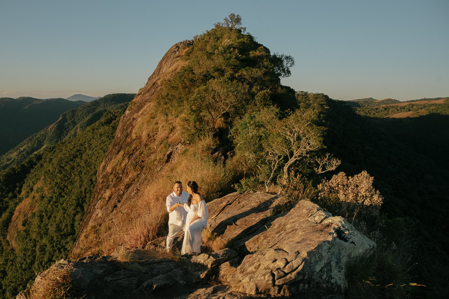 Juliano ajuda Letícia a caminhar sobre a crista rochosa da Pedra do Baú. Ela veste um vestido branco fluido e ele camisa branca. O cenário destaca a formação rochosa imponente sob a luz dourada do fim de tarde na Serra da Mantiqueira.