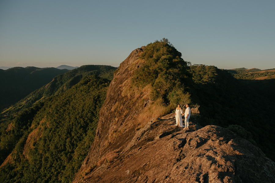 Letícia e Juliano estão de mãos dadas no topo de uma crista rochosa na Pedra do Baú. Ela usa um vestido branco fluido e ele camisa branca. O casal é emoldurado pela imensidão das montanhas da Serra da Mantiqueira sob a luz dourada do pôr do sol.