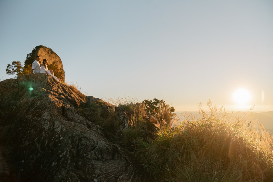 Letícia e Juliano estão sentados no topo de uma formação rochosa, observando o pôr do sol na Serra da Mantiqueira. O sol radiante no horizonte cria um brilho dourado sobre a vegetação e destaca a silhueta do casal contra o céu claro.