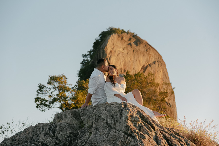 Juliano beija a testa de Letícia enquanto estão sentados em uma rocha alta na Pedra do Baú. Ela sorri de olhos fechados. O sol do fim de tarde ilumina o casal e a imponente formação rochosa ao fundo contra um céu azul limpo.
