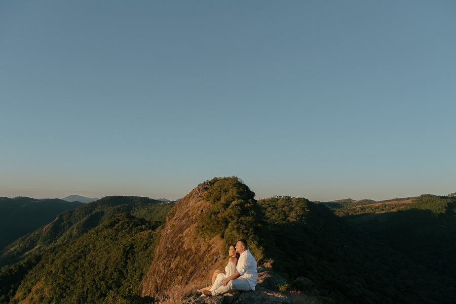 Letícia e Juliano estão sentados no topo da Pedra do Baú, observando a imensidão das montanhas sob a luz dourada do pôr do sol. Ela veste um vestido branco fluido e ele camisa branca, emoldurados pelo cenário da Serra da Mantiqueira.