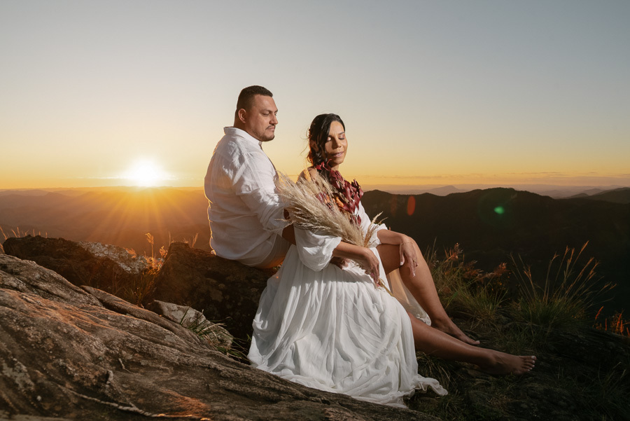 Leticia e Juliano sentados em uma rocha na Pedra do Baú durante o pôr do sol. Ela segura um buquê de capim seco e veste vestido branco fluido; ele usa camisa branca. O sol no horizonte cria um brilho dourado intenso sobre as montanhas.