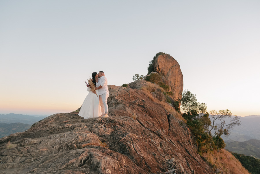 Leticia e Juliano se beijam no topo de uma crista rochosa na Pedra do Baú durante o pôr do sol. Ela segura um buquê de flores secas e veste vestido branco fluido. Ao fundo, a formação rochosa imponente sob o céu em tons de degradê.