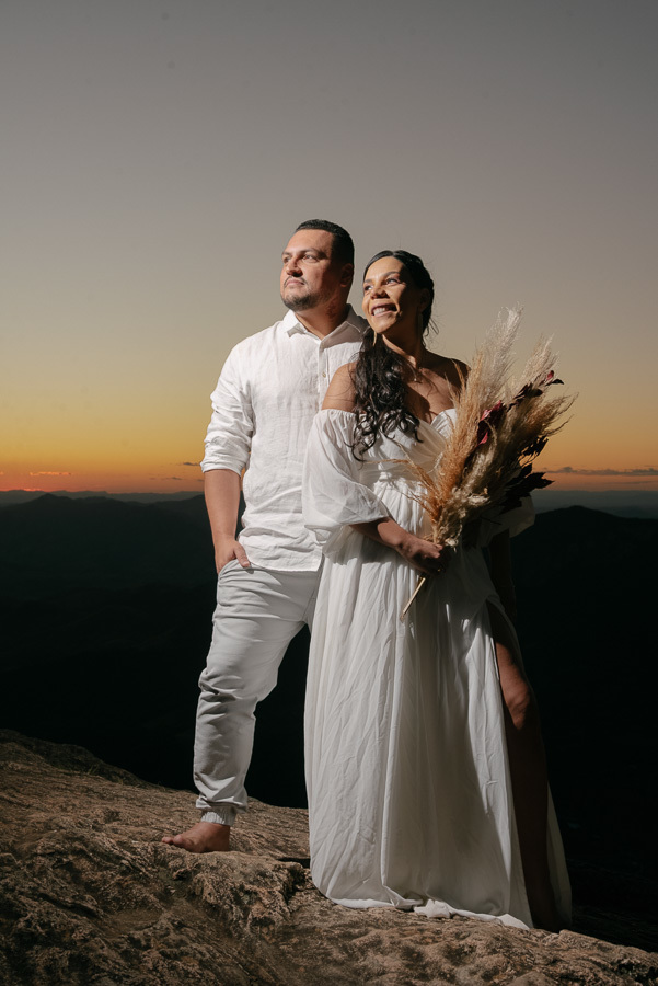 Juliano e Letícia posam no topo da Pedra do Baú durante o crepúsculo. Ele veste camisa branca e ela um vestido branco fluido, segurando um buquê de capim seco. Ao fundo, o céu exibe um degradê de laranja e azul sobre as silhuetas das montanhas.