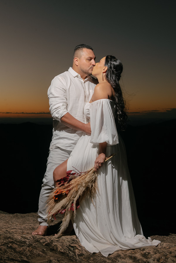 Juliano e Letícia se beijam no topo da Pedra do Baú durante o crepúsculo. Ele veste camisa branca e ela um vestido branco fluido, segurando um buquê de capim seco. O casal é iluminado contra um céu escurecido com um horizonte laranja vibrante.