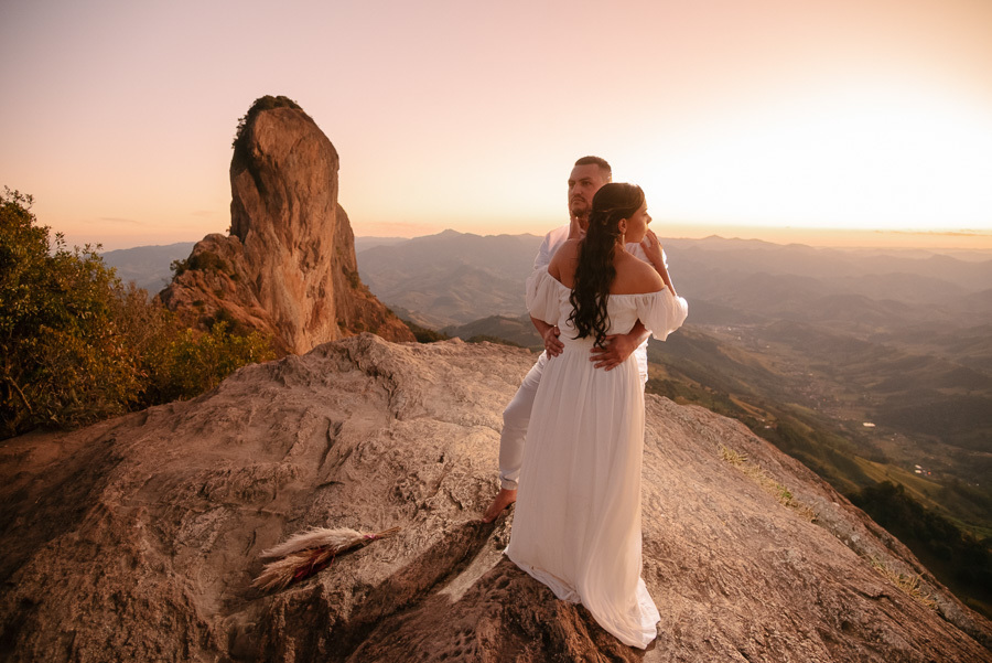 Leticia e Juliano abraçados no topo da Pedra do Baú durante o pôr do sol. Ela veste vestido branco e ele camisa clara. Ao fundo, a imponente crista rochosa e as montanhas da Serra da Mantiqueira sob uma luz dourada e céu alaranjado.
