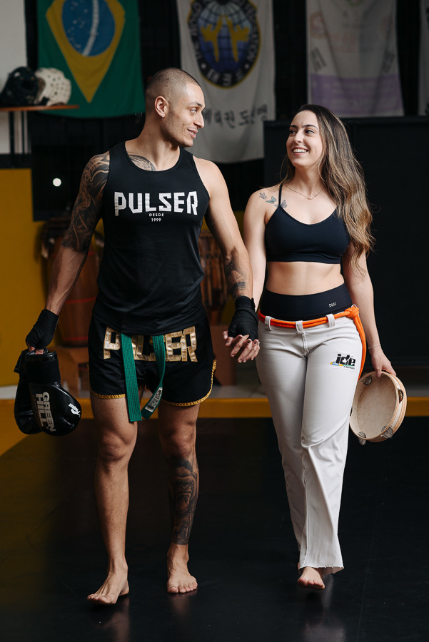 Luidgi e Aline caminham sorridentes no ensaio pré-casamento, unindo kickboxing e capoeira. Fotografia de casal lifestyle com luvas e pandeiro na academia. Registro profissional e autêntico para inspirar noivos que buscam fotos criativas e únicas.