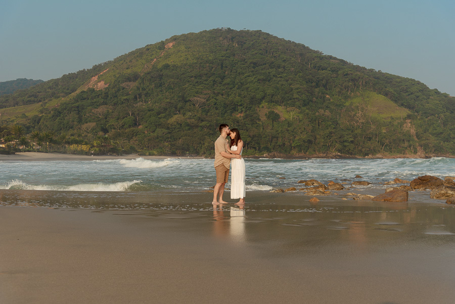 Foto de Matthaus e Rebeca no ensaio pré-casamento na Praia de Camburizinho, São Sebastião. Fotógrafo profissional captura momento romântico do casal com beijo na testa à beira-mar e montanhas ao fundo. Sentimento e conexão na areia.