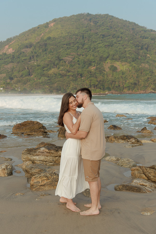 Matthaus e Rebeca em momento carinhoso no ensaio pré-casamento na Praia de Camburizinho, São Sebastião. Fotografia profissional destacando o sorriso da noiva e as pedras da praia, com o mar e a natureza de fundo.