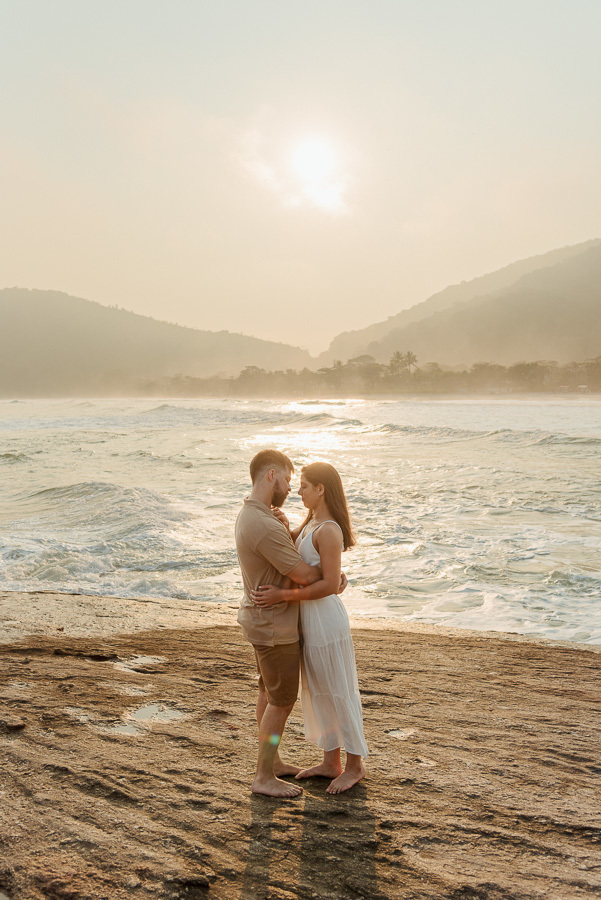 Matthaus e Rebeca abraçados em uma rocha plana durante ensaio pré-casamento na Praia de Camburizinho, São Sebastião. Luz dourada e ondas do mar compõem o cenário profissional de fotografia de casal.