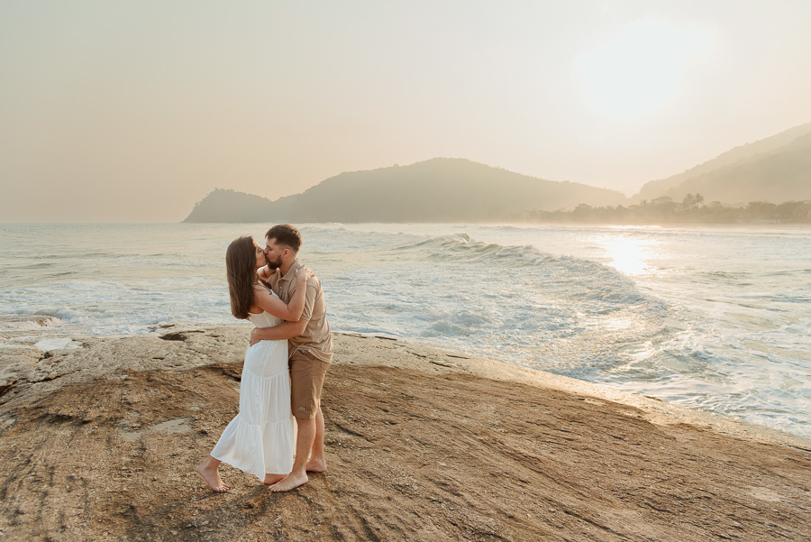 Registro profissional do beijo de Matthaus e Rebeca no ensaio pré-casamento na Praia de Camburizinho. Composição ampla destacando a imensidão do mar de São Sebastião e a sintonia do casal sobre as pedras.