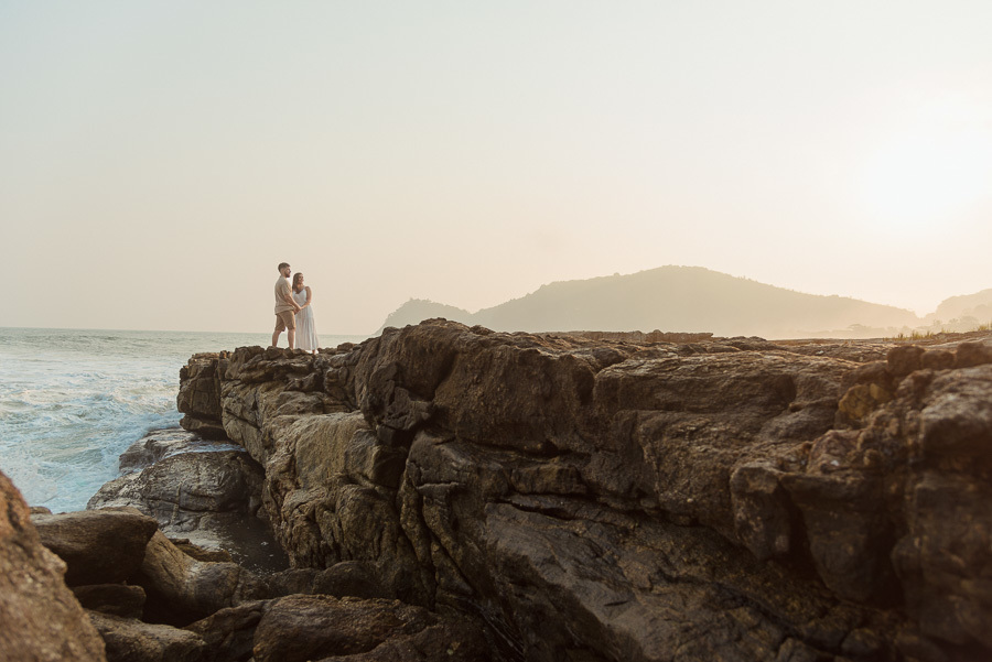 Foto profissional de Matthaus e Rebeca no topo de grandes rochas na Praia de Camburizinho. O ensaio pré-casamento em São Sebastião destaca a imensidão das falésias e o mar agitado sob a luz suave do entardecer no litoral norte.