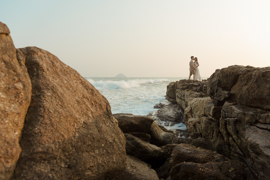 Composição fotográfica de Matthaus e Rebeca se beijando à distância sobre rochas imponentes na Praia de Camburizinho. Ensaio pré-casamento em São Sebastião que valoriza a perspectiva, a força da natureza e o amor do casal.