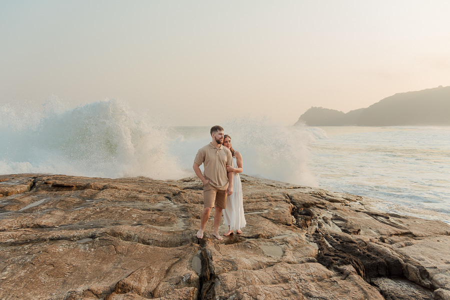 Foto profissional de Matthaus e Rebeca no ensaio pré-casamento em Camburizinho, São Sebastião. O casal posa sobre as rochas enquanto uma onda estoura ao fundo, criando um cenário dinâmico e impactante no litoral.