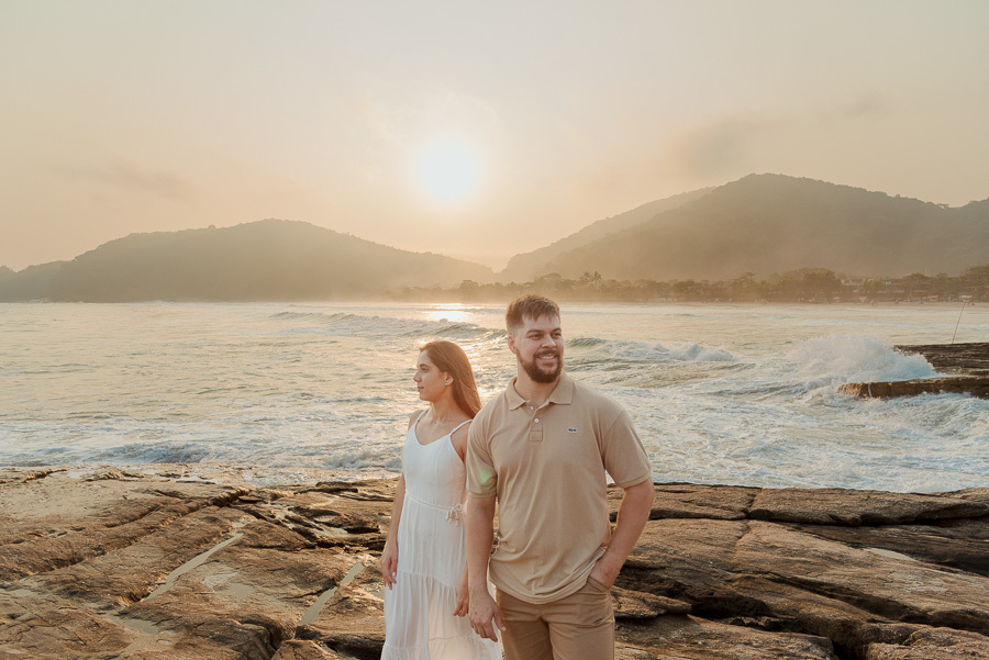 Fotografia profissional de casal em Camburizinho, São Sebastião. Matthaus e Rebeca posam sorridentes sobre as pedras durante o ensaio pré-casamento, com o mar, montanhas e o sol poente criando um cenário iluminado e romântico.