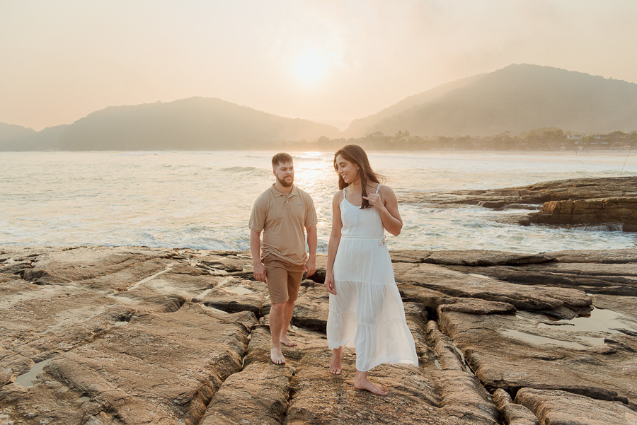 Fotografia profissional de Matthaus e Rebeca caminhando pelas rochas da Praia de Camburizinho, em São Sebastião. O ensaio pré-casamento captura a descontração do casal sob a luz dourada do pôr do sol com o mar e as montanhas ao fundo.