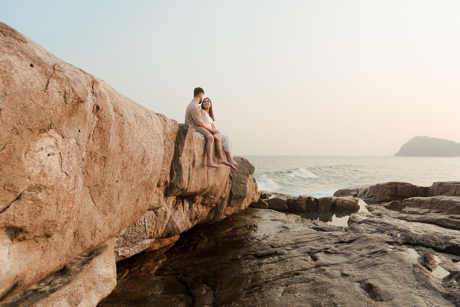 Matthaus levanta Rebeca em um abraço romântico sobre as rochas da Praia de Camburizinho. Fotografia de pré-casamento em São Sebastião capturada durante o pôr do sol, destacando a alegria e a sintonia do casal.