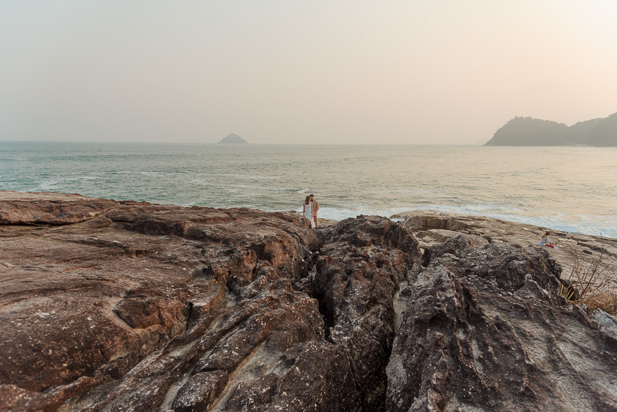 Matthaus e Rebeca caminham descontraidamente sobre as rochas da Praia de Camburizinho. Registro espontâneo de ensaio pré-casamento em São Sebastião sob a luz dourada do entardecer paulista.