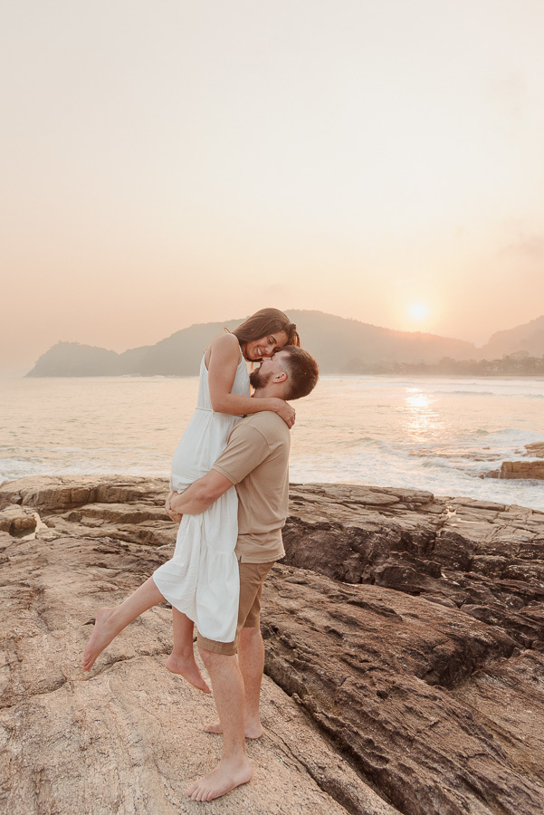 Matthaus levanta Rebeca em um abraço romântico sobre as rochas da Praia de Camburizinho. Fotografia de pré-casamento em São Sebastião capturada durante o pôr do sol, destacando a alegria e a sintonia do casal.