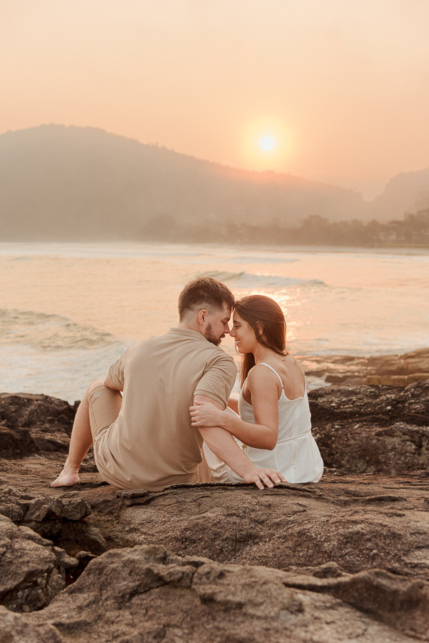 Matthaus e Rebeca sentados de costas observando o pôr do sol na Praia de Camburizinho. Final de ensaio pré-casamento em São Sebastião com tons quentes e atmosfera de serenidade.