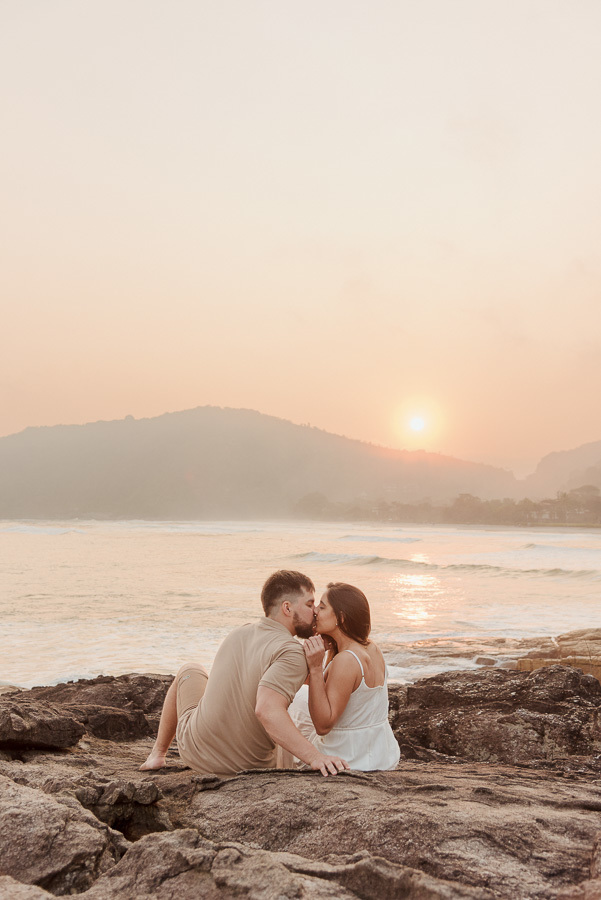 Beijo romântico de Matthaus e Rebeca sentados nas rochas durante o pôr do sol na Praia de Camburizinho. Fotografia profissional de pré-casamento em São Sebastião com tons quentes, mar calmo e silhueta das montanhas ao fundo.