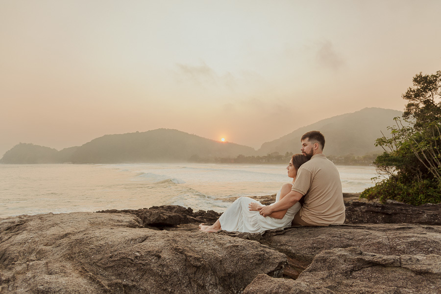Matthaus e Rebeca sentados de costas observando o pôr do sol na Praia de Camburizinho. Final de ensaio pré-casamento em São Sebastião com tons quentes e atmosfera de serenidade.