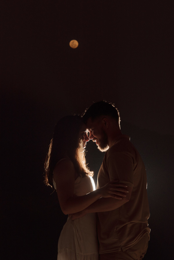 Fotografia artística noturna de Matthaus e Rebeca em silhueta durante ensaio pré-casamento. O casal aparece unido sob o luar na Praia de Camburizinho, com iluminação de contorno (rim light) destacando a conexão romântica em São Sebastião.