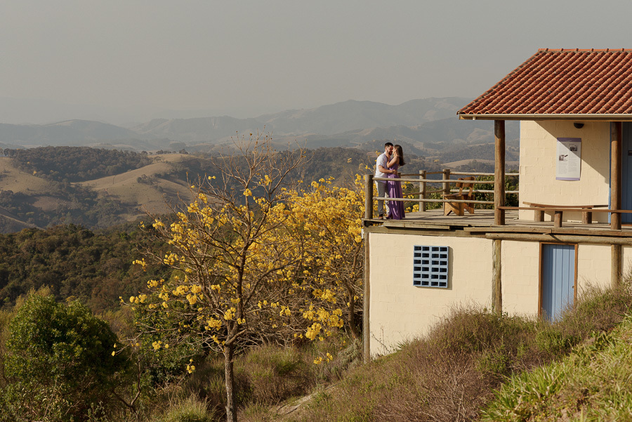 Matthaus e Rebeca abraçados no deck de madeira do Lavandário de Cunha. Fotografia de ensaio pré-casamento que destaca a beleza do Ipê-amarelo e as colinas da Serra da Mantiqueira. Registro profissional e romântico no interior de São Paulo.