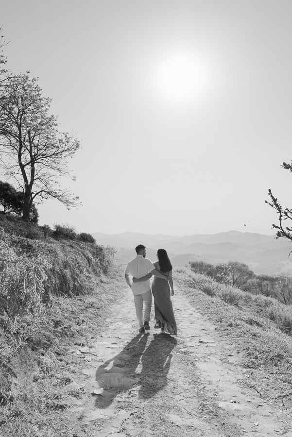 Matthaus e Rebeca caminham abraçados sob o sol no Lavandário de Cunha. Fotografia pre-wedding em preto e branco com silhueta e luz intensa, simbolizando a jornada do casal. Registro profissional e artístico para um álbum de noivado na serra.