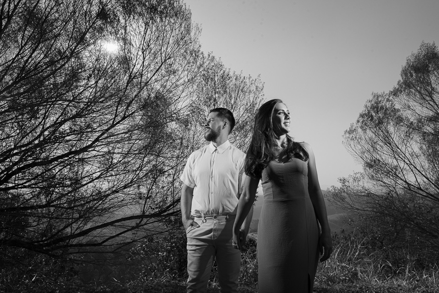Matthaus e Rebeca sorriem para o futuro no Lavandário de Cunha. Fotografia pre-wedding em preto e branco com luz dramática entre as árvores da Serra da Mantiqueira. Registro profissional e artístico que celebra o amor e a jornada do casal.