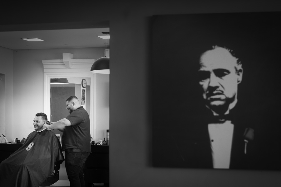 Foto em preto e branco do dia do casamento de Letícia e Juliano no Troina. Em um plano médio, o noivo sorri enquanto o barbeiro finaliza seu corte de cabelo. À direita, em primeiro plano, destaca-se um quadro de Marlon Brando como 