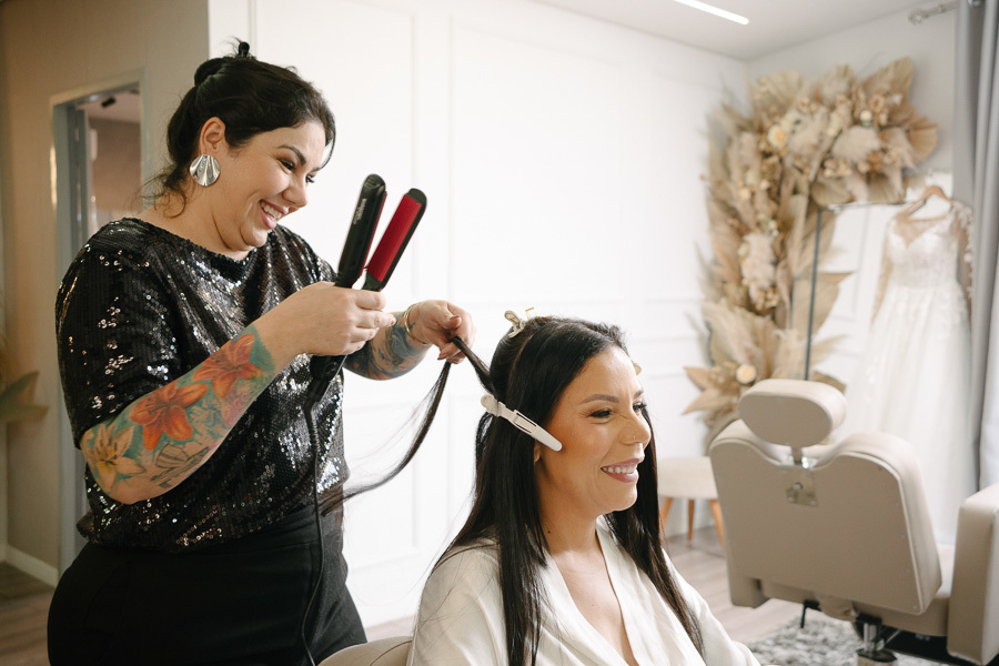 Preparação da noiva Letícia para o casamento em São José dos Campos. Ela sorri enquanto a cabeleireira usa uma prancha no seu cabelo. Ao fundo, detalhes da decoração com flores secas e o vestido de noiva pendurado completam o cenário emocionante.