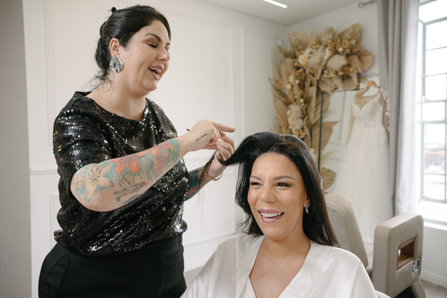 A noiva Letícia sorri alegremente enquanto a profissional finaliza seu penteado para o casamento. Ao fundo, o vestido de noiva branco está pendurado em um cabide, cercado por um arranjo de flores secas em tons terrosos, criando um clima de sonho.