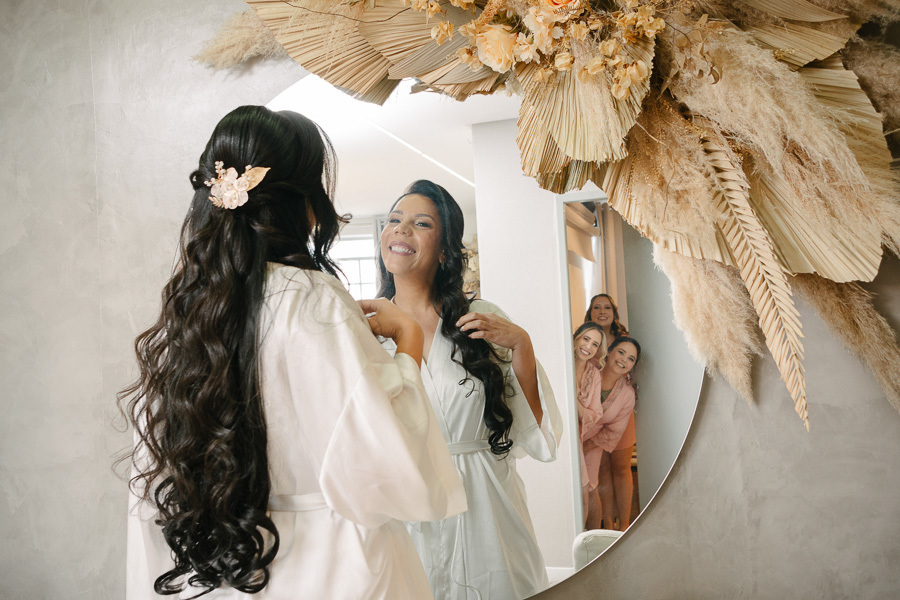 A noiva Letícia admira seu penteado semipreso com acessório floral no espelho redondo, decorado com arranjo de flores secas. No reflexo, ela sorri enquanto suas madrinhas aparecem ao fundo da porta, compartilhando a emoção do making of no Troina.