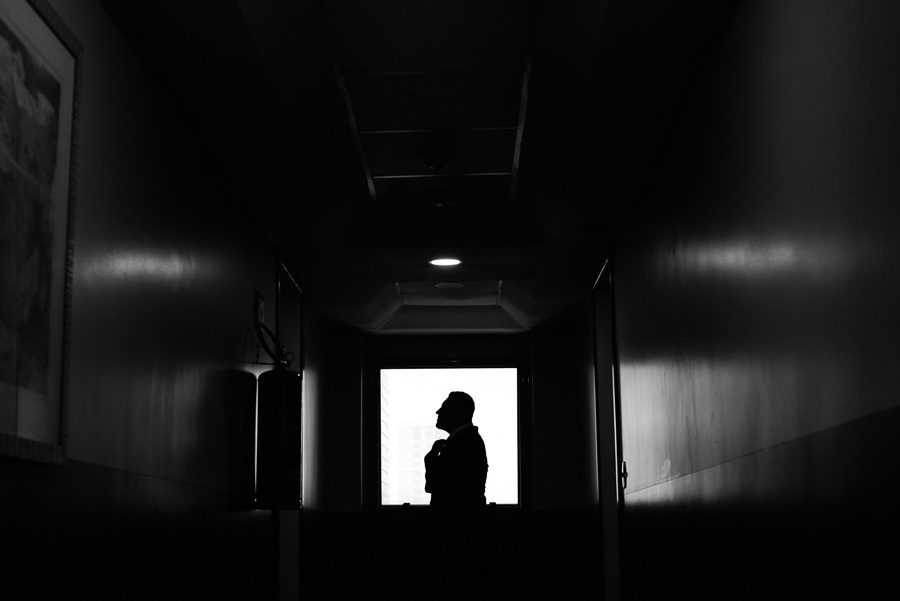 Silhueta em preto e branco do noivo Juliano em um corredor escuro, posicionado contra a luz de uma janela ao fundo enquanto ajusta o terno. A imagem artística captura um momento de reflexão e preparação antes do casamento inesquecível no Troina.