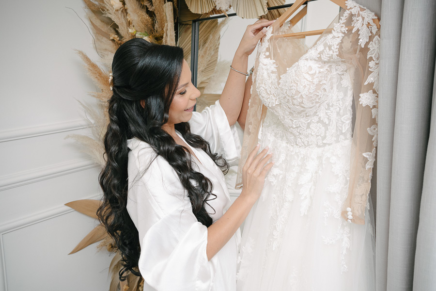 A noiva Letícia, vestindo um robe de seda branco, toca com admiração seu vestido de noiva pendurado. O vestido possui mangas longas em renda floral detalhada. Ao fundo, um arranjo de flores secas completa a cena do making of no Troina.