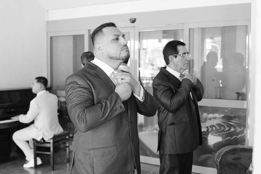 Foto em preto e branco do noivo Juliano ajustando a gravata antes do casamento no Troina. Ao seu lado, um senhor realiza o mesmo gesto, enquanto ao fundo um músico toca piano. O momento captura a elegância e a expectativa para a cerimônia inesquecível.