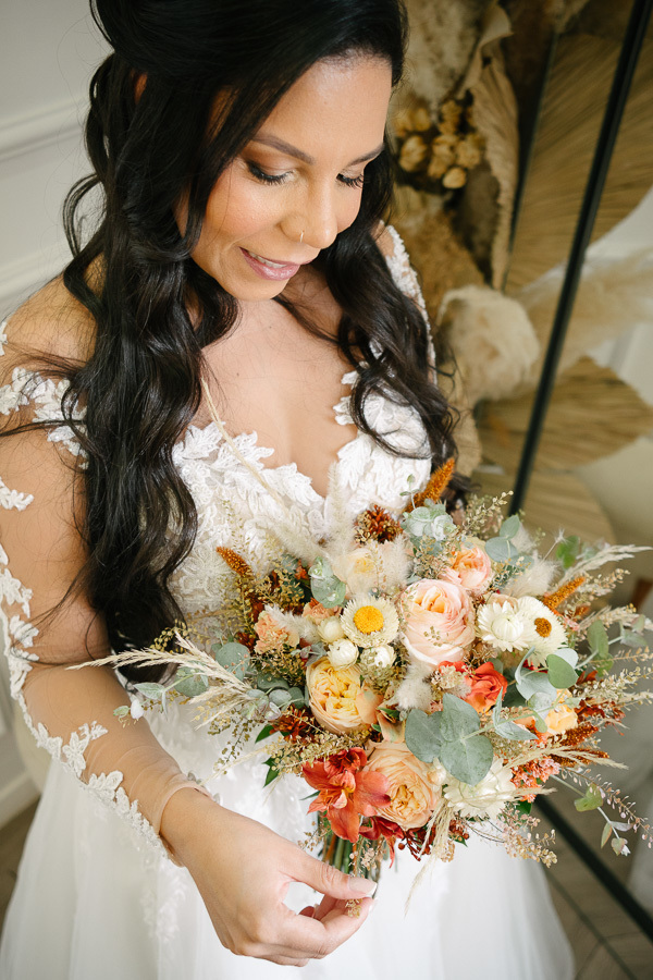 Close-up da noiva Letícia admirando seu buquê de flores naturais e secas em tons de pêssego, amarelo e terracota. Ela veste o vestido de noiva rendado, evidenciando a emoção e os detalhes delicados do seu casamento inesquecível no Troina.