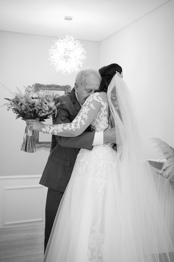 Foto em preto e branco da noiva Letícia abraçando seu pai de forma emocionante no Troina. Ela segura o buquê enquanto o pai fecha os olhos, transmitindo um momento de puro sentimento e amor familiar antes da cerimônia de casamento em São José dos Campos.