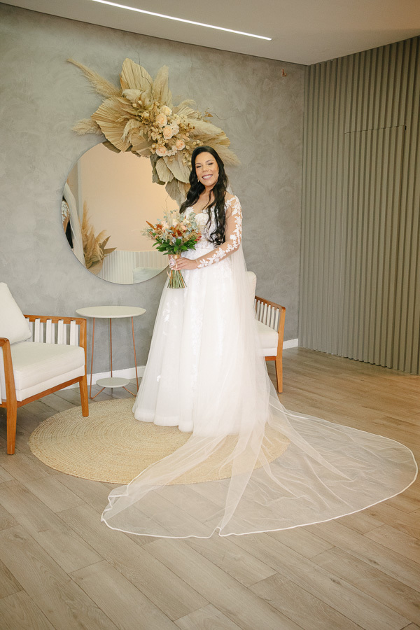 A noiva Letícia posa sorridente, já pronta com seu vestido rendado de mangas longas e véu longo. Ela segura o buquê colorido no cenário do Troina, em São José dos Campos, em frente a um espelho redondo com arranjo de flores secas e capim-dos-pampas.