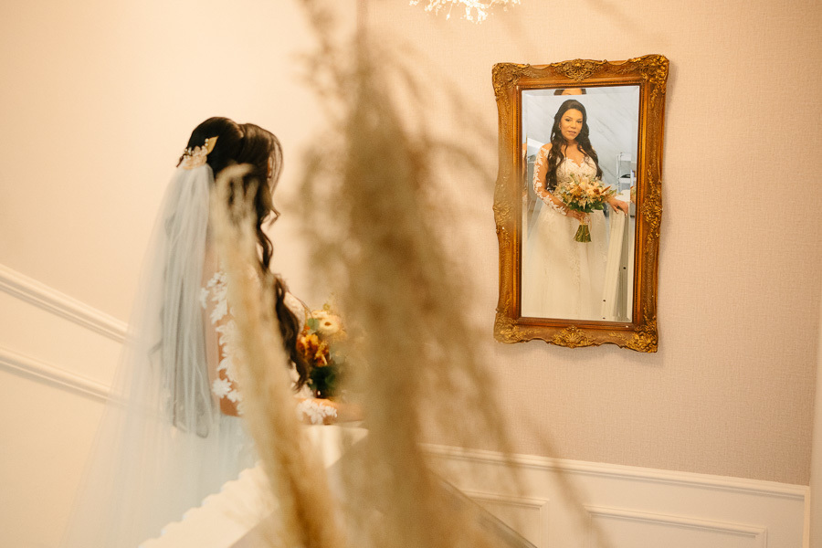 Reflexo da noiva Letícia em um espelho de moldura dourada vintage. Ela está pronta, segurando o buquê e vestindo seu vestido rendado com véu. A imagem, com foco artístico entre folhagens, captura a elegância final do seu making of no Troina.