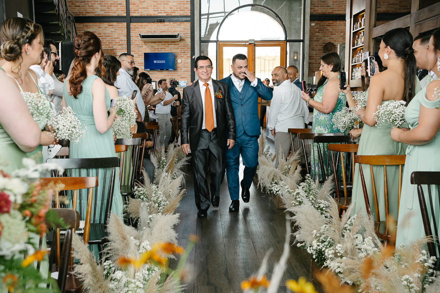 O noivo Juliano entra emocionado na cerimônia no Troina, secando uma lágrima enquanto caminha pelo corredor decorado com capim-dos-pampas. Ele está acompanhado por um senhor, sob os olhares de madrinhas de verde e convidados em São José dos Campos.