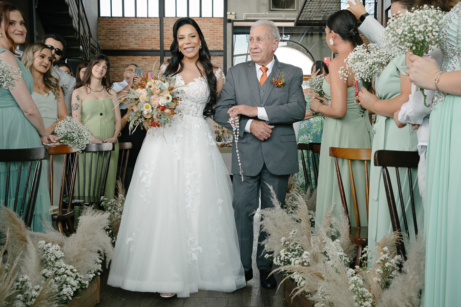 A noiva Letícia entra radiante na cerimônia no Troina, de braço dado com seu pai, que segura um terço. Ela carrega seu buquê colorido enquanto caminha pelo corredor decorado com capim-dos-pampas, sob os olhares atentos de madrinhas e convidados.