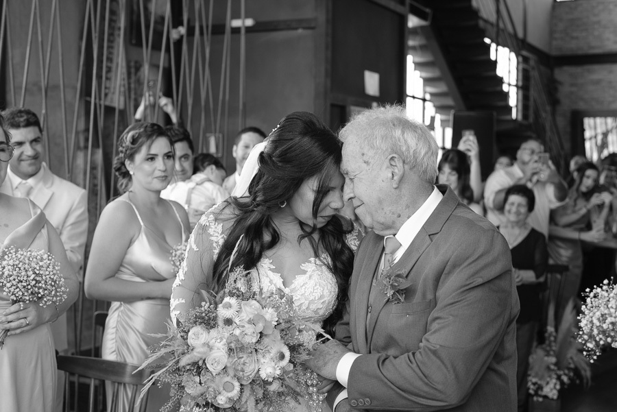 Foto em preto e branco da noiva Letícia encostando a testa na do seu pai em um momento de carinho no altar. Ela segura o buquê enquanto ele a entrega ao noivo. Ao fundo, convidados e madrinhas observam a cena emocionante da cerimônia no Troina.