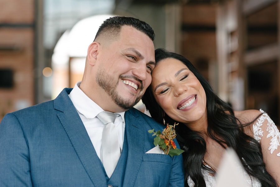 Close-up dos noivos Letícia e Juliano sorrindo no altar do Troina. A noiva encosta a cabeça carinhosamente no ombro do noivo, que veste um terno azul. A imagem transmite a felicidade e a intensidade do amor celebrada em São José dos Campos.